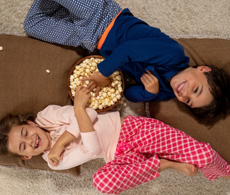 Blije kinderen, jongen + meisje, in hun pyjama's op de bank met een bak popcorn.